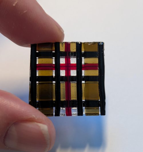 May include: A small, square decorative piece with a plaid pattern. The design features black vertical and horizontal lines, with gold and red accents. The piece is held between two fingers.